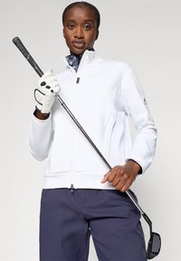 Hvit golfjakke med glidelås og stående krage, teksturerte ermer, kombinert med marineblå bukser. Modellen holder en golfkølle i en hanskekledd hånd.