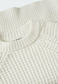 Gros plan d’un pull blanc en maille montrant un col rond côtelé et un tissu à motif gaufré texturé.