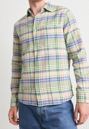 Mand iført en langærmet skjorte med beige bundfarve og tern i blå, grøn og rød, kombineret med blå jeans.