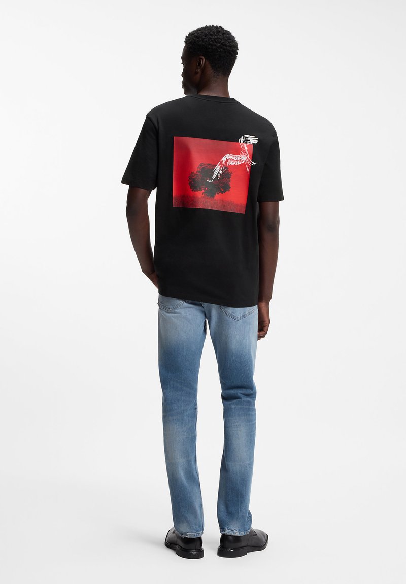 Άνδρας που φοράει μαύρο μπλουζάκι με κόκκινο τετράγωνο και λευκό σχέδιο πουλιού στην πλάτη, συνδυασμένο με ανοιχτό μπλε τζιν και μαύρα παπούτσια.