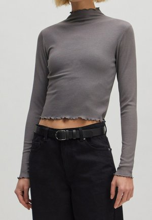 Persona che indossa un crop top a maniche lunghe grigio a coste con bordi a lattuga e pantaloni neri a vita alta con una cintura nera.