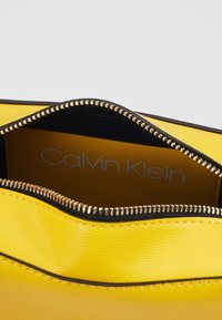 Borsa gialla in pelle con una texture liscia, dettagli con cerniera nera e logo interno. Il design presenta un bordo superiore curvo.