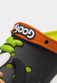 Μαύρο λαστιχένιο παπούτσι τύπου κλογκ με πορτοκαλί λουράκι που γράφει «Goofy» σε λευκό, με διακόσμηση καρτούν χαρακτήρα σε πράσινο και άσπρο στο μπροστινό μέρος.