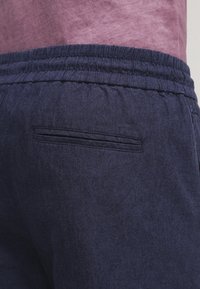 Marineblauwe linnen broek met een elastische tailleband en geplooide textuur, en een discreet achterzakje. Gladde stof met een zachte afwerking.