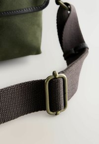Dunkelbrauner verstellbarer Stoffgurt mit Messingschnalle, befestigt an einer grünen Tragetasche auf weißem Hintergrund.