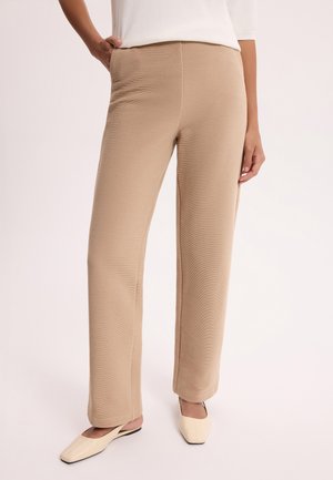 Beżowe spodnie o prostych nogawkach z prążkowanej tkaniny, noszone z kremowymi balerinami na niskim obcasie z kwadratowym noskiem oraz białą bluzką, przedstawione na stojącej osobie.