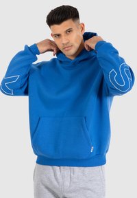 Blauwe pullover hoodie van een zachte stof met een kangoeroezak, voorzien van witte grafische accenten op de mouwen en een trekkoordcapuchon.