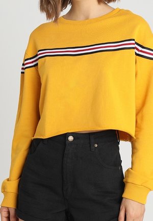 Persona con un jersey corto amarillo con rayas rojas, blancas y negras en el pecho y pantalones cortos negros de talle alto.