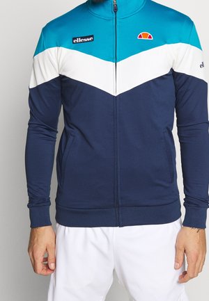 Mann trägt eine marineblaue, weiße und türkisfarbene Sportjacke mit Reißverschluss und ellesse-Logos, kombiniert mit weißen Hosen vor einfarbigem Hintergrund.