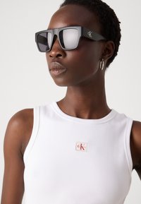 Occhiali da sole neri oversize con una finitura lucida, dotati di lenti piatte e di un logo CK sul lato. Il modello indossa un top a canotta bianco a costine.