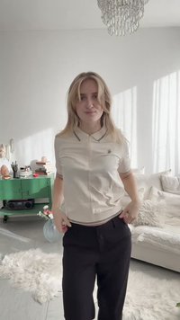 Νέα γυναίκα με μπεζ πόλο και μαύρο παντελόνι στέκεται σε φωτεινό σαλόνι με λευκό καναπέ, πράσινη ντουλάπα, πολυέλαιο και ηλιακό φως στους τοίχους.