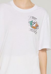 Biała bawełniana koszulka z nadrukowanym logo w zielonym, pomarańczowym i czarnym kolorze z napisem "TRUE RELIGION" na lewym piersi.