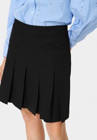Jupe plissée noire avec une texture lisse et une taille élastique, associée à une chemise bleu clair à rayures. La longueur de la jupe arrive au-dessus du genou.