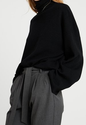 Femme portant un pull à col roulé noir ample rentré dans un pantalon paperbag gris taille haute avec une ceinture en tissu.