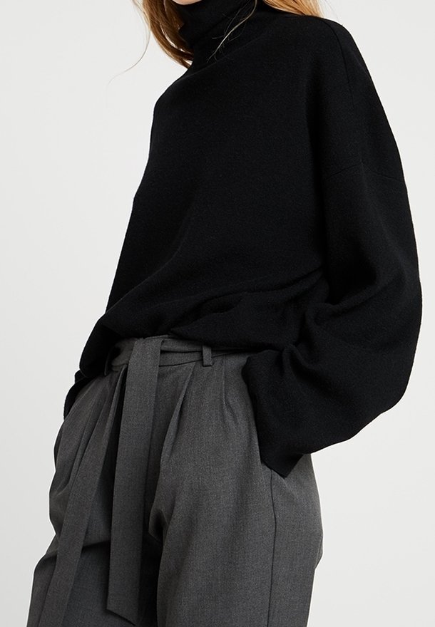 Femme portant un pull à col roulé noir ample rentré dans un pantalon paperbag gris taille haute avec une ceinture en tissu.