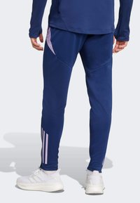 Pantalons de sport navy avec des accents latéraux violets, présentant un design ajusté et un tissu texturé. Associés à des chaussures de sport blanches.