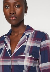 Marineblauw en scharlakenrood geruite pyjama-top met witte piping, voorzien van een borstzak en een gekantelde kraag. Zacht katoenen stof, casual ontwerp.