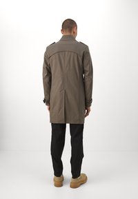 Trench coat verde oliva scuro con colletto alto, spalline sulle spalle e vestibilità sartoriale, abbinato a pantaloni neri e calzature color cammello.