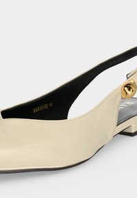 Μπεζ δερμάτινο παπούτσι slingback με χαμηλό τακούνι και χρυσή αγκράφα, που δείχνει πλάγια και μερική εσωτερική όψη.