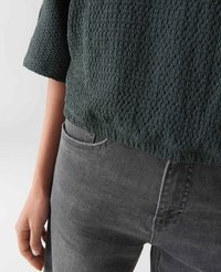 Persoon in een donkergroen, gestructureerd, kort shirt dat in hooggetailleerde grijze jeans is gestoken, met de hand ontspannen naast zich.