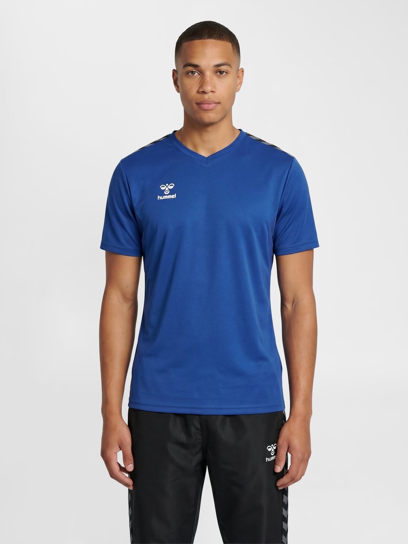 Ung mand iført en blå hummel sportstrøje og sorte hummel træningsbukser, stående mod en ensartet lys baggrund.