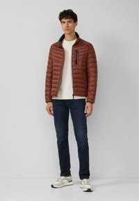 Bruine pufferjack met hoge kraag, horizontaal gestikte patronen, ritssluiting aan de voorkant en een zwarte borstzak, gecombineerd met donkere jeans en sneakers.