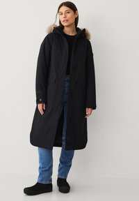 Giacca parka nera lunga con cappuccio foderato in pelliccia, dotata di chiusura con zip e bottoni, tasche laterali e taglio dritto. Indossata sopra jeans e scarpe nere.