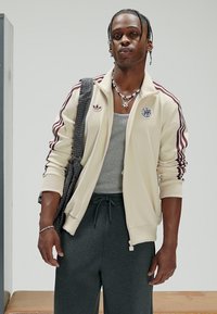 Casaco bege com fecho éclair, riscas vermelhas e logótipo, camisola interior cinzenta, calças de treino cinzento escuro, e uma mala aos quadrados sobre o ombro, a posar numa superfície clara.