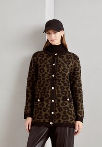 Leopardprint cardigan in donker groen en bruin, met een knopenflap, ribbels aan de onderkant en twee voorzakken. Draagt over een zwarte coltrui.