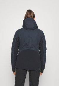 Veste à capuche bleu foncé au design ajusté. Comprend des sections de tissu contrastées et une texture lisse. La vue de dos montre une coupe mi-longue.
