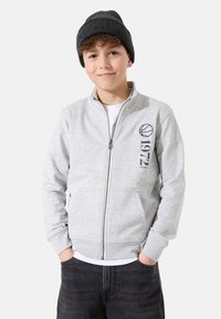 Sudadera gris con cremallera y cuello acanalado, con bolsillos laterales y un gráfico de baloncesto con "1972" impreso. Lleva puesta una gorra de punto negra.