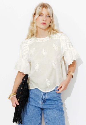 Femme blonde portant un chemisier en dentelle blanche avec des manches bouffantes, un jean bleu et des bracelets dorés, tenant une pochette noire à franges.