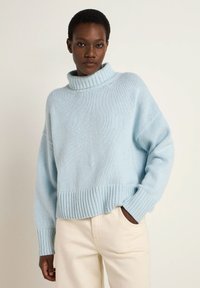 Light blue, oversized knit sweater with a high ribbed collar and ribbed hem. Soft texture and loose fit. Paired with cream-colored pants.