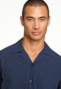 Marineblauw overhemd met korte mouwen en een gestructureerde afwerking, twee voorkwijdte zakken en een puntige kraag. Heeft subtiele stiksels en een knoopsluiting.
