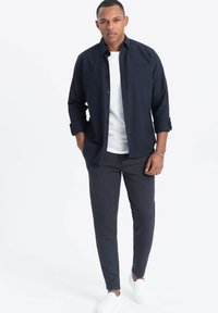 Camicia blu con bottoni sopra una maglietta bianca, abbinata a pantaloni scuri a fantasia e scarpe bianche senza lacci. Il modello è in piedi contro uno sfondo neutro.