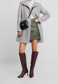 Grau-er Wollmantel, grüner Leder-Minirock und braune knielange Absatzstiefel; schwarze Schultertasche mit Krokodilstruktur und goldenen Beschlägen.