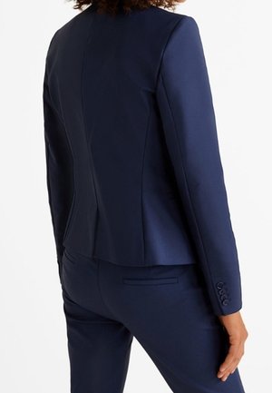 Nainen pukeutunut istuvaan laivastonsiniseen bleiseriin ja siihen sopiviin housuihin, kuvattuna takaa, käsi rentona sivulla.