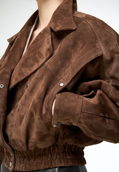 Veste en daim marron avec boutons-pression, taille élastique, portée par une personne avec une main dans une poche.