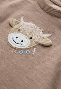 Brown fabric t-shirt featuring a cream and tan embroidered cow face with a smiling expression and the word "Moo!" in pastel colors.