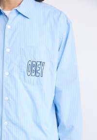 Camisa de botón a rayas azul claro con un bolsillo en el pecho que lleva la palabra bordada "OBEY". La tela parece suave y ligera.