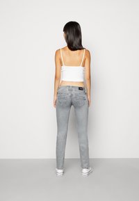 Mujer de pie de espaldas, vistiendo un top corto blanco, jeans gris claro y zapatillas blancas, contra un fondo claro y liso.