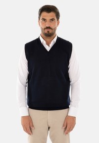 Gilet in maglia senza maniche di colore blu navy, con scollatura a V, indossato sopra una camicia bianca a maniche lunghe. Presenta una texture liscia e una forma semplice.