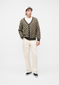 Νέος άνδρας που φοράει μπεζ και σκούρο καφέ καρό ζακέτα, λευκό πουκάμισο, κρεμ παντελόνι, λευκό καπέλο και μαύρα αθλητικά παπούτσια, στέκεται μπροστά σε λευκό φόντο.