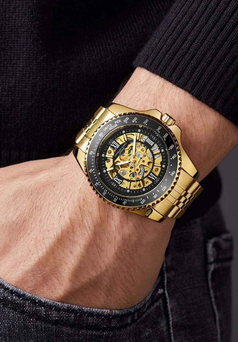 Gouden roestvrijstalen horloge met een zwarte wijzerplaat, voorzien van een skeletonontwerp, Romeinse cijfers en een gestructureerde band. Zichtbare details van de kroon en de lunette.