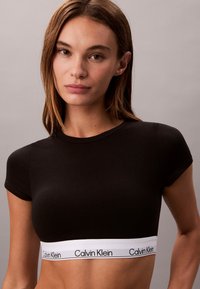 Kvinde med glat brunt hår iført en sort crop top med et hvidt Calvin Klein-logo bånd, stående mod en ensfarvet grå baggrund.