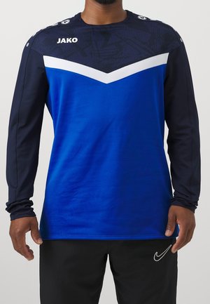 Uomo che indossa una maglia sportiva a maniche lunghe con motivo a chevron nei colori navy, blu e bianco e pantaloni neri con logo Nike bianco.