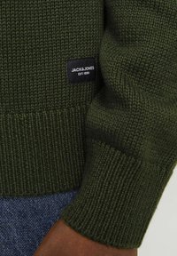 Pull vert en tricot avec des poignets côtelés, arborant une étiquette noire avec l'inscription "JACK & JONES EST. 1990" brodée sur le côté gauche.