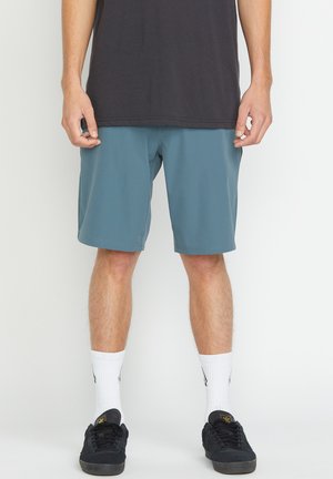 Shorts teal pour homme avec une texture lisse, ceinture élastique et ourlets cousus, associés à des baskets noires et des chaussettes blanches chevilles.