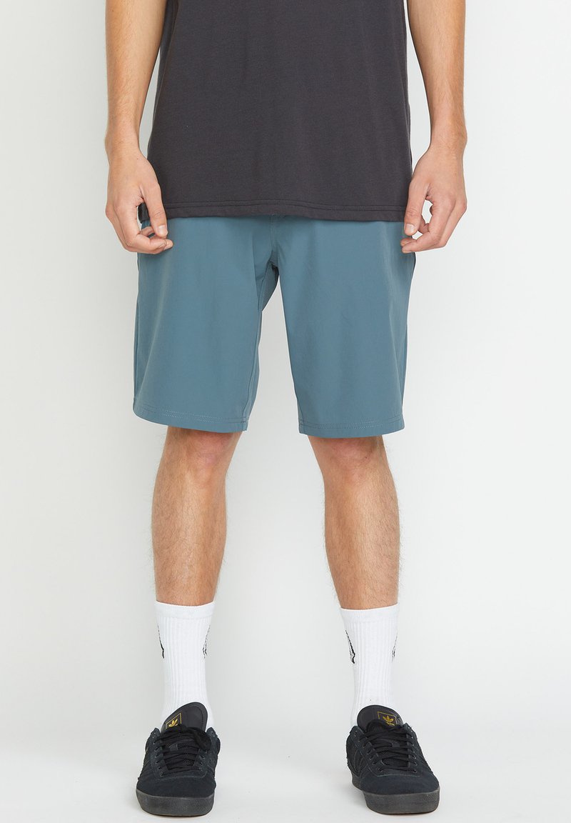 Shorts teal pour homme avec une texture lisse, ceinture élastique et ourlets cousus, associés à des baskets noires et des chaussettes blanches chevilles.