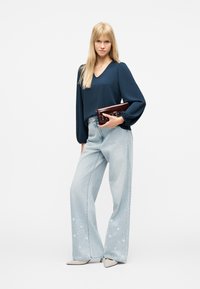 Γυναίκα με μπλε μπλούζα μακριού μανικιού και ανοιχτό μπλε τζιν παντελόνι με φαρδιά μπατζάκια και λευκή κεντητή λουλουδένια διακόσμηση, κρατάει καφέ σκούρο φάκελο, στέκεται σε λευκές γόβες.